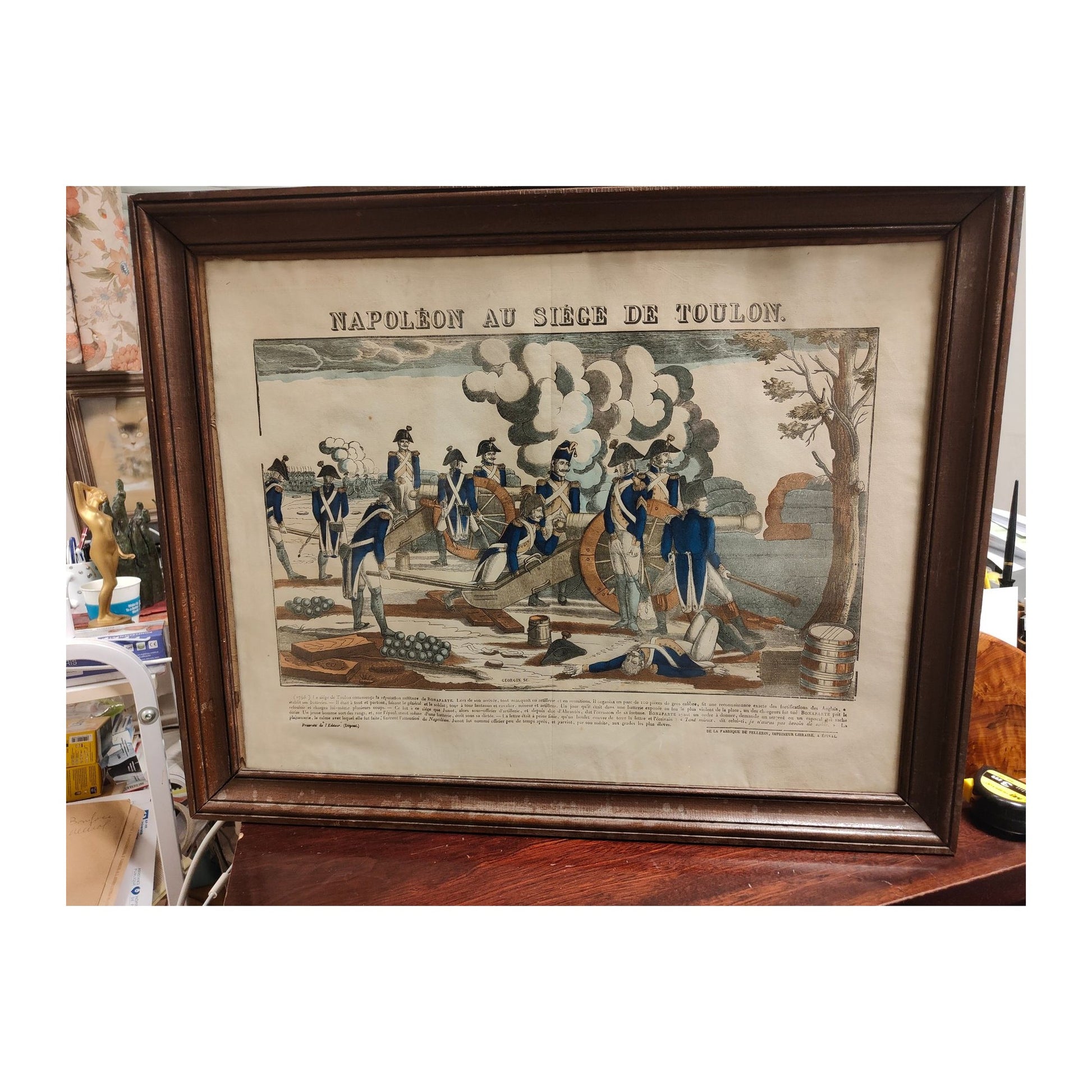 Gravure ancienne en couleurs représentant Napoléon au siège de Toulon, imagerie Pellerin d’Épinal XIXe siècle, estampe encadrée sous verre avec pliure au centre.
